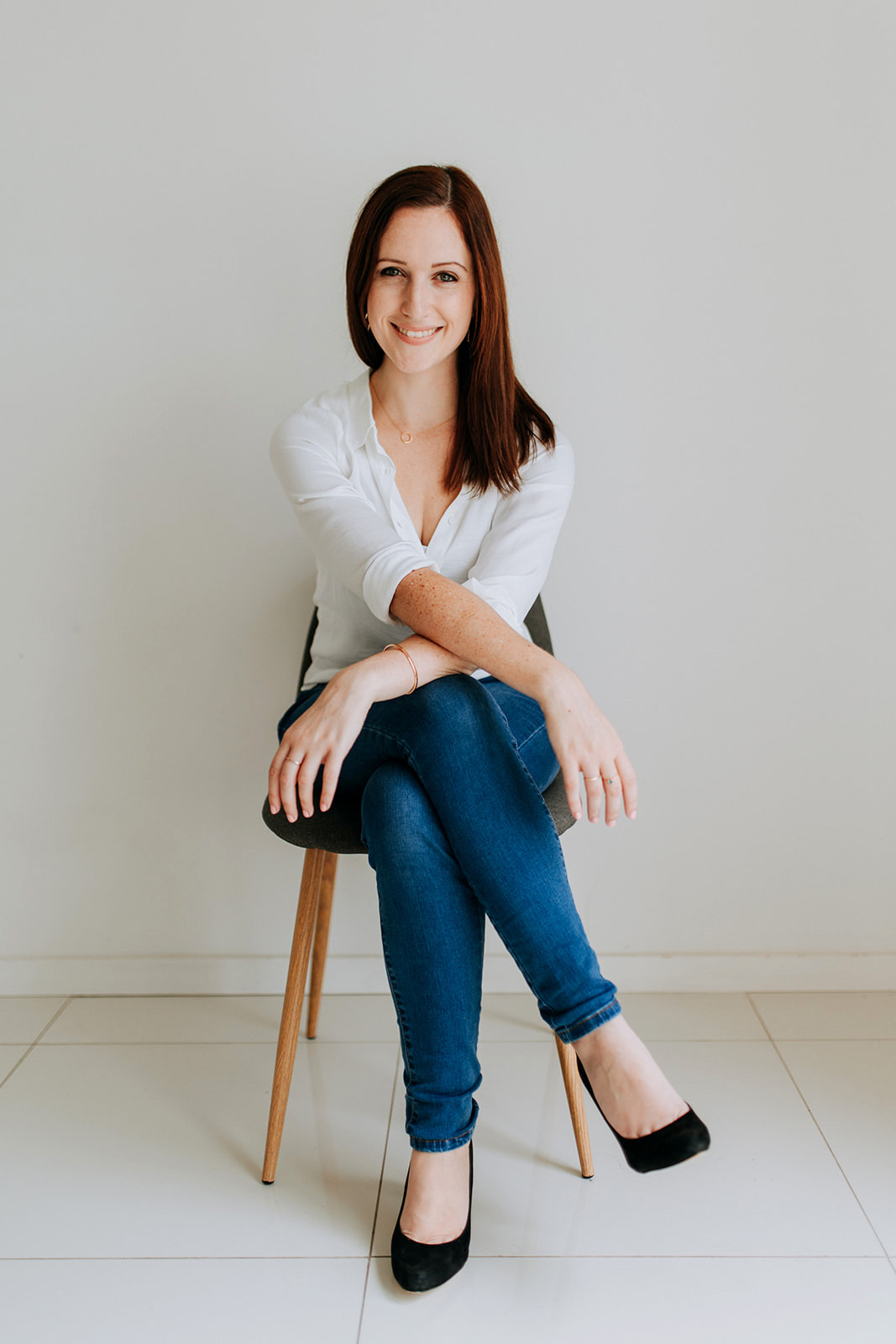A photo of Ally. She is a white woman in her early thirties with brown hair. She is wearing a white blouse and blue jeans. She is sitting on a chair against a plain white wall. She is smiling.